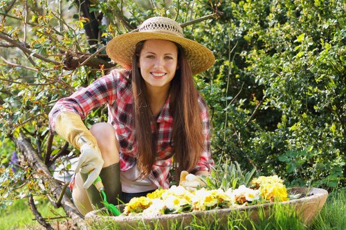 A young woman with long brown hair, wearing a straw gardening hat, a red and white checkered shirt, and gardening gloves, is crouching in a lush garden. She is smiling and working on a flower bed that contains a variety of yellow and white flowers. Behind her, there are leafy green shrubs and trees, with some branches extending into the foreground. The garden features a well-maintained lawn, organic soil areas, and wooden garden borders, indicating outdoor care and landscaping work typical of a domestic garden in the Queens Park area, with natural sunlight illuminating the scene and creating a vibrant, healthy outdoor environment. This setting exemplifies professional gardening and lawn care services offered by Lawn Mowing Queens Park, focusing on maintaining lush lawns, flower beds, and garden borders that thrive in a suburban outdoor space.