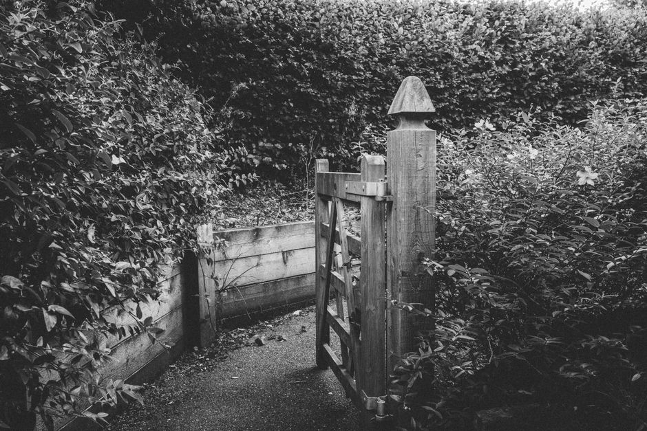 A narrow garden pathway with a simple wooden gate featuring a pointed cap on its post, situated between dense bushes and trees. The pathway is paved with small gravel or pebbles, bordered by a weather