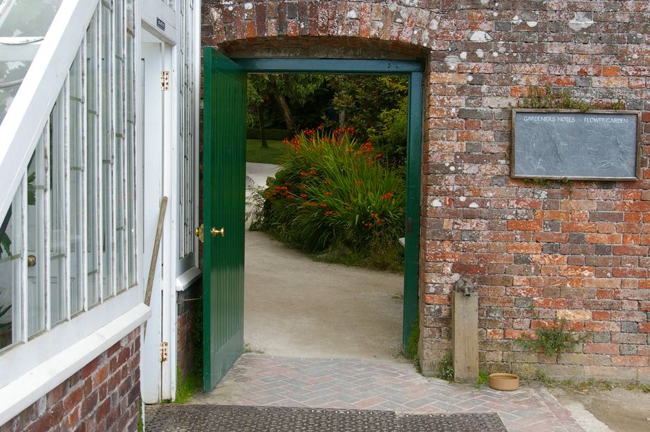 A narrow garden pathway with a simple wooden gate featuring a pointed cap on its post, situated between dense bushes and trees. The pathway is paved with small gravel or pebbles, bordered by a weather